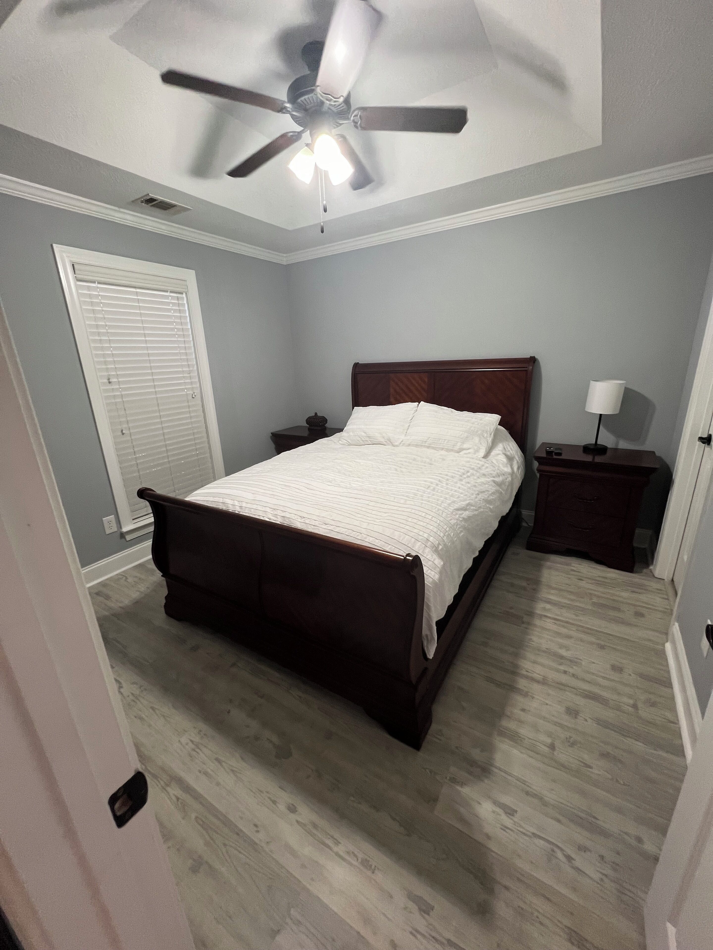 Quarto com cama de madeira, mesa de cabeceira, luminária, ventilador de teto e janela com persianas.