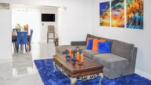 Een woonkamer met een grijze bank, blauwe en oranje kussens, een houten salontafel met kaarsen, en een blauw vloerkleed.