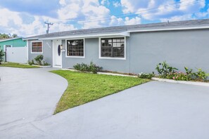 A single-story building with a grey exterior, a small front yard with grass and flowers, and a driveway leading to the entrance. - Beautiful 4-bedroom house in Heart of  Miami Gardens with POOL, AC and WiFi (Miami Gardens)