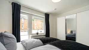 Et soveværelse med seng, gardiner ved vinduet, et sengebord med lampe og et spejl på væggen.