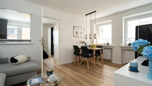 En moderne lejlighed med stue, spiseplads og køkken. Der er en sofa, et sofabord i glas og et spisebord med stole.
