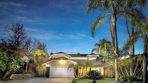 A two-story house with a garage, surrounded by palm trees and a well-maintained lawn. - Serene hillside oasis with pool, spa, & BBQ - 20 min to beach. 31-night minimum. (Oceanside)