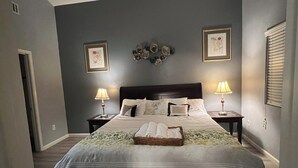 Un dormitorio con una cama grande, dos mesitas de noche con lámparas, una obra de arte en la pared y una puerta a la izquierda.