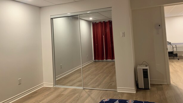 A room with a sliding glass door leading to a bathroom, a white air conditioning unit, and a wooden floor. - Spacious basement in nice area (Salaberry-de-Valleyfield)