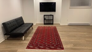 A living room with a black sofa, a red patterned rug, a flat-screen TV, and a window with blinds. - Spacious basement in nice area (Salaberry-de-Valleyfield)