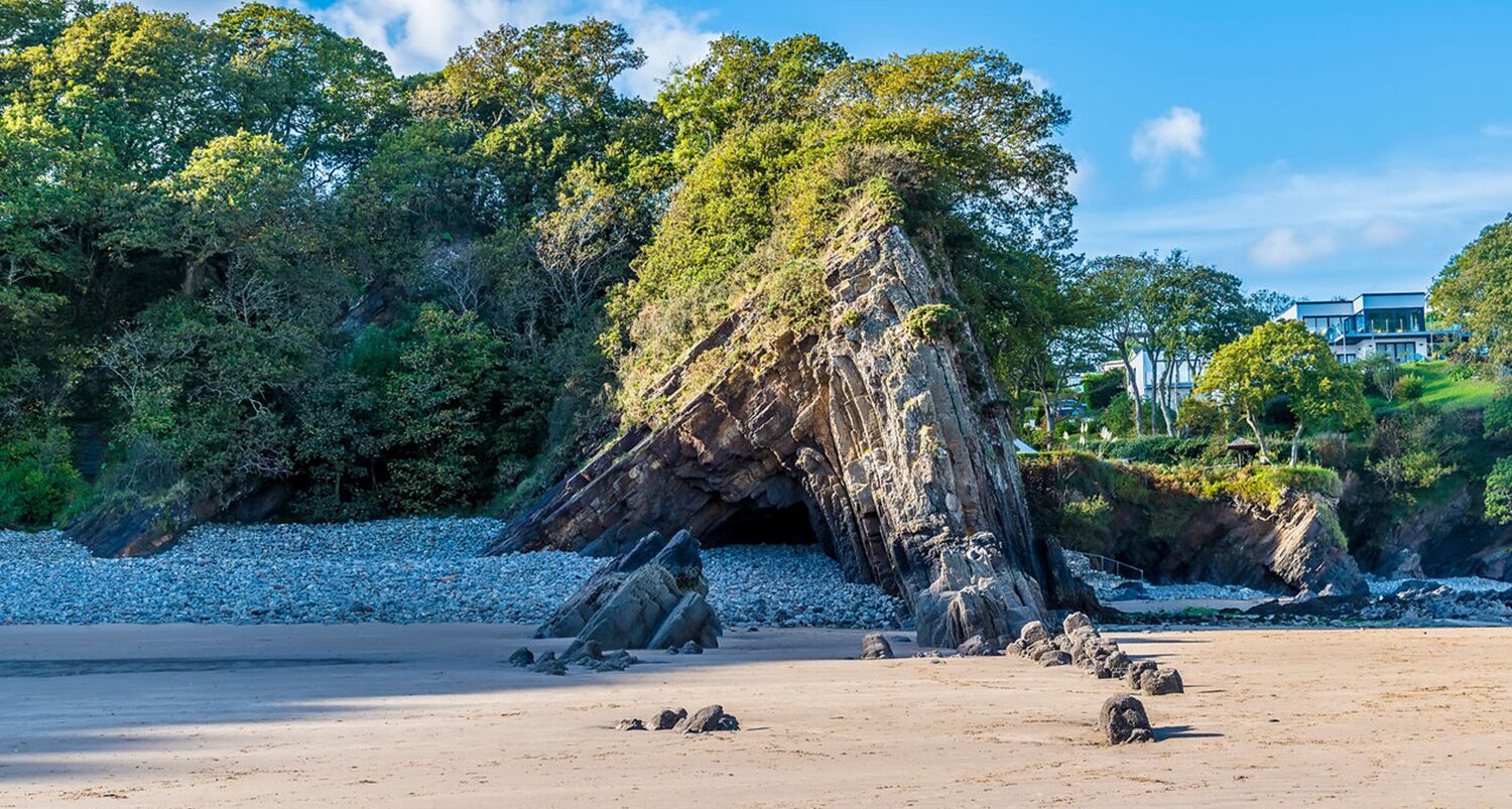 A natural rock formation with a cave entrance, surrounded by trees and a beach.