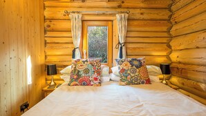 A wooden cabin bedroom with a bed, two pillows, a window with curtains, bedside lamps, and a view of trees outside. - Pwll Cabin, Holiday Home Marros, Sleeps 2, 1 bedrooms,  bathrooms (Carmarthen)