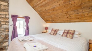 A bedroom with a sloped ceiling, a stone wall, a bed with a white duvet and plaid pillows, a nightstand with a lamp, and a window with purple curtains. - Pwll Cottage, Holiday Cottage Marros, Sleeps 6, 3 bedrooms,  bathrooms (Carmarthen)