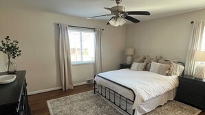 Une chambre à coucher comprenant un lit, un ventilateur de plafond, une table de chevet, une commode et une fenêtre avec des rideaux.