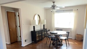 Une pièce spacieuse dotée d’un plancher en bois, d’une table à manger avec des chaises, d’un ventilateur de plafond et d’une grande fenêtre avec des rideaux.