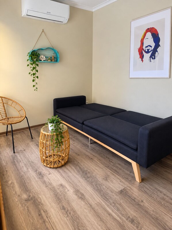 Una habitación con un sofá oscuro, una silla de mimbre, una mesa redonda de mimbre con una planta y un aire acondicionado instalado en la pared.