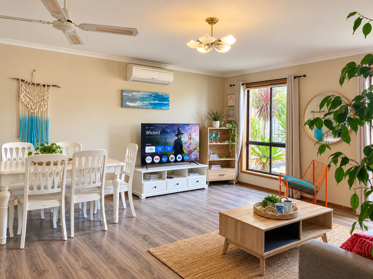 A living room with a flat-screen TV, white furniture, a ceiling fan, and a large window with a view of greenery.