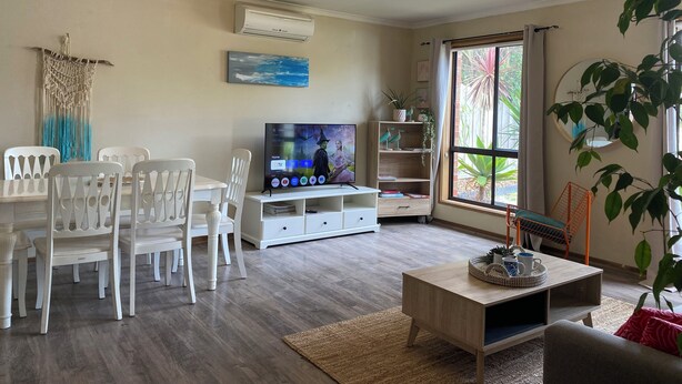 A living room with a dining area, a TV, a ceiling fan, and a large window with a view of greenery.