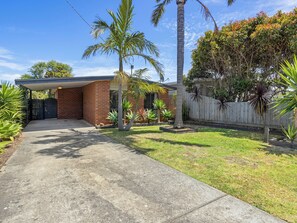 A residential property with a paved driveway leading to a brick house, surrounded by a wooden fence and various tropical plants.