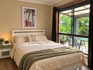 A bedroom with a bed, a nightstand, a lamp, a window with curtains, and a view of greenery outside.