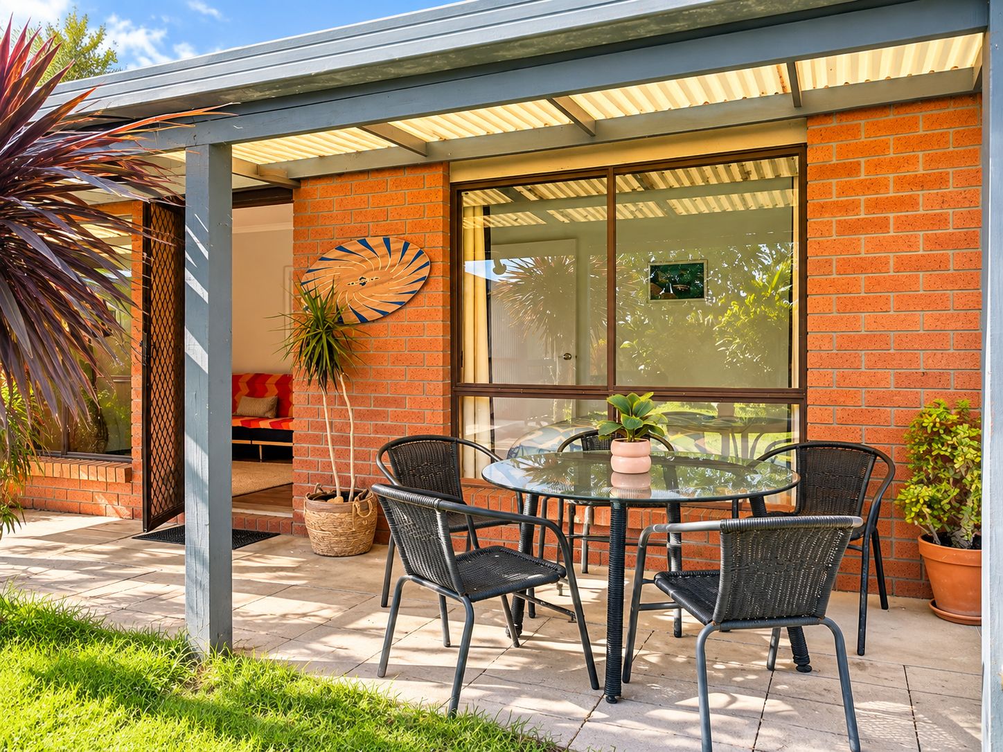 An outdoor patio with a glass table and four chairs, surrounded by a brick wall and a glass door.