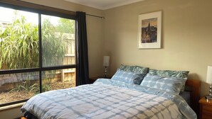 A bedroom with a large window, a bed with patterned bedding, a cityscape painting, and a nightstand with a lamp.