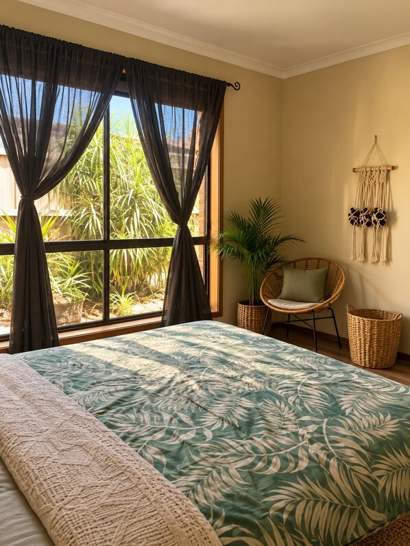 A bedroom with a large bed, a wicker chair, a macrame wall hanging, and a view of greenery outside.