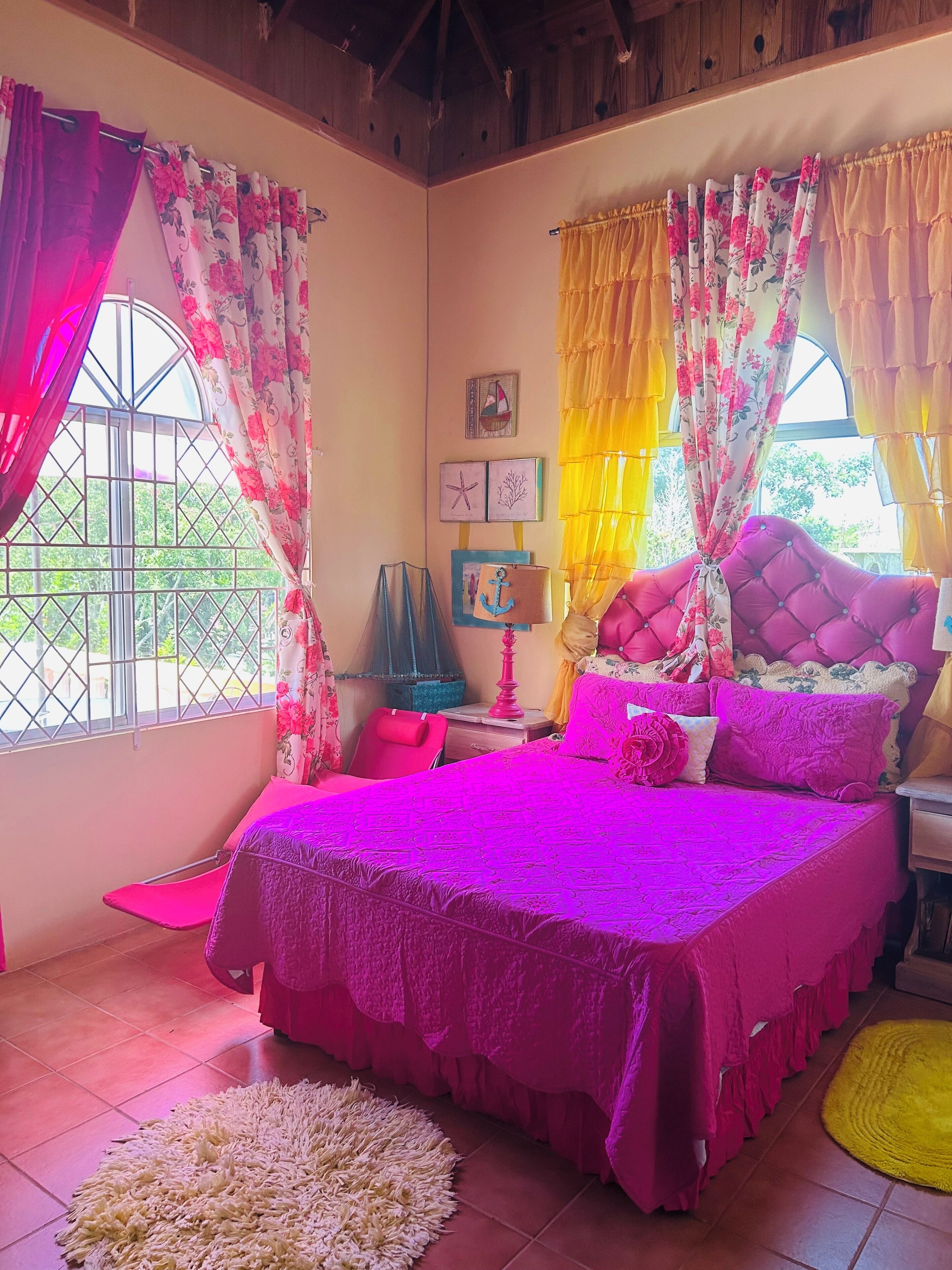 A bedroom with a large bed, curtains, a nightstand, a lamp, and a window with a view of greenery.