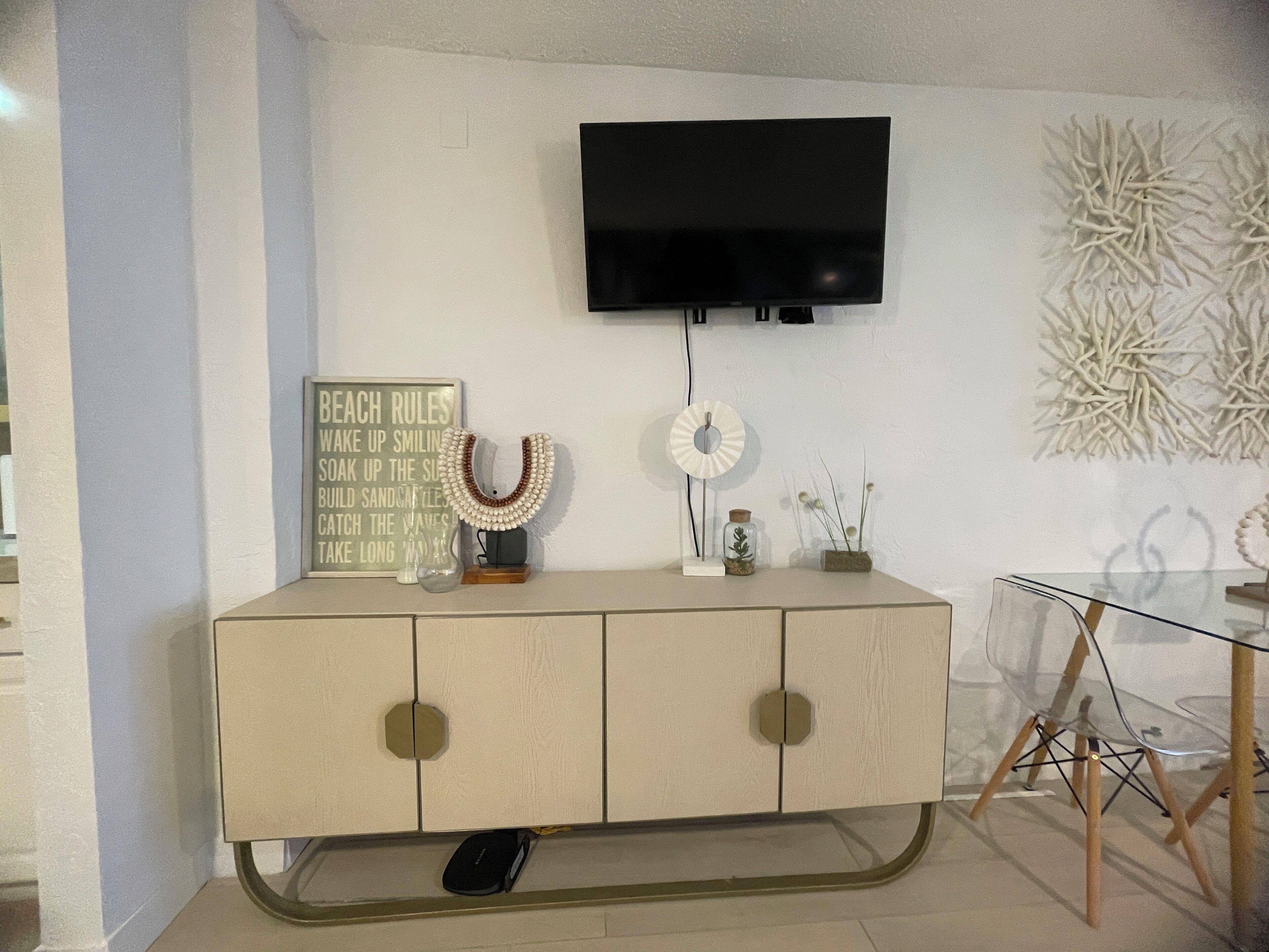 A modern living room with a flat-screen TV mounted on the wall, a decorative wall piece, a framed sign, and a glass table with chairs.