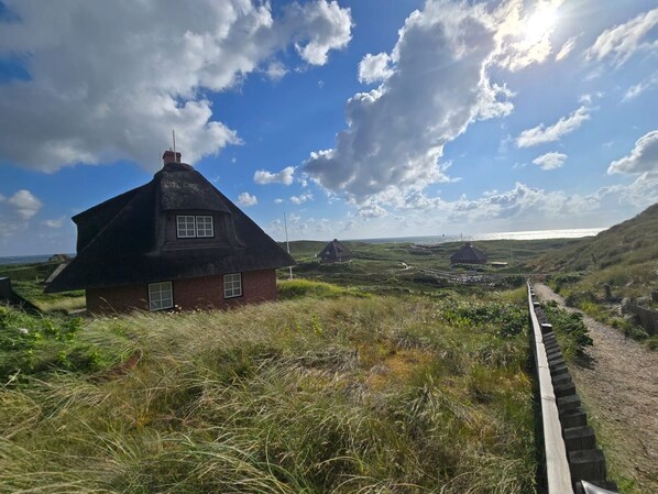 A traditional thatched-roof house with a brick base, surrounded by grassy fields and a clear sky.