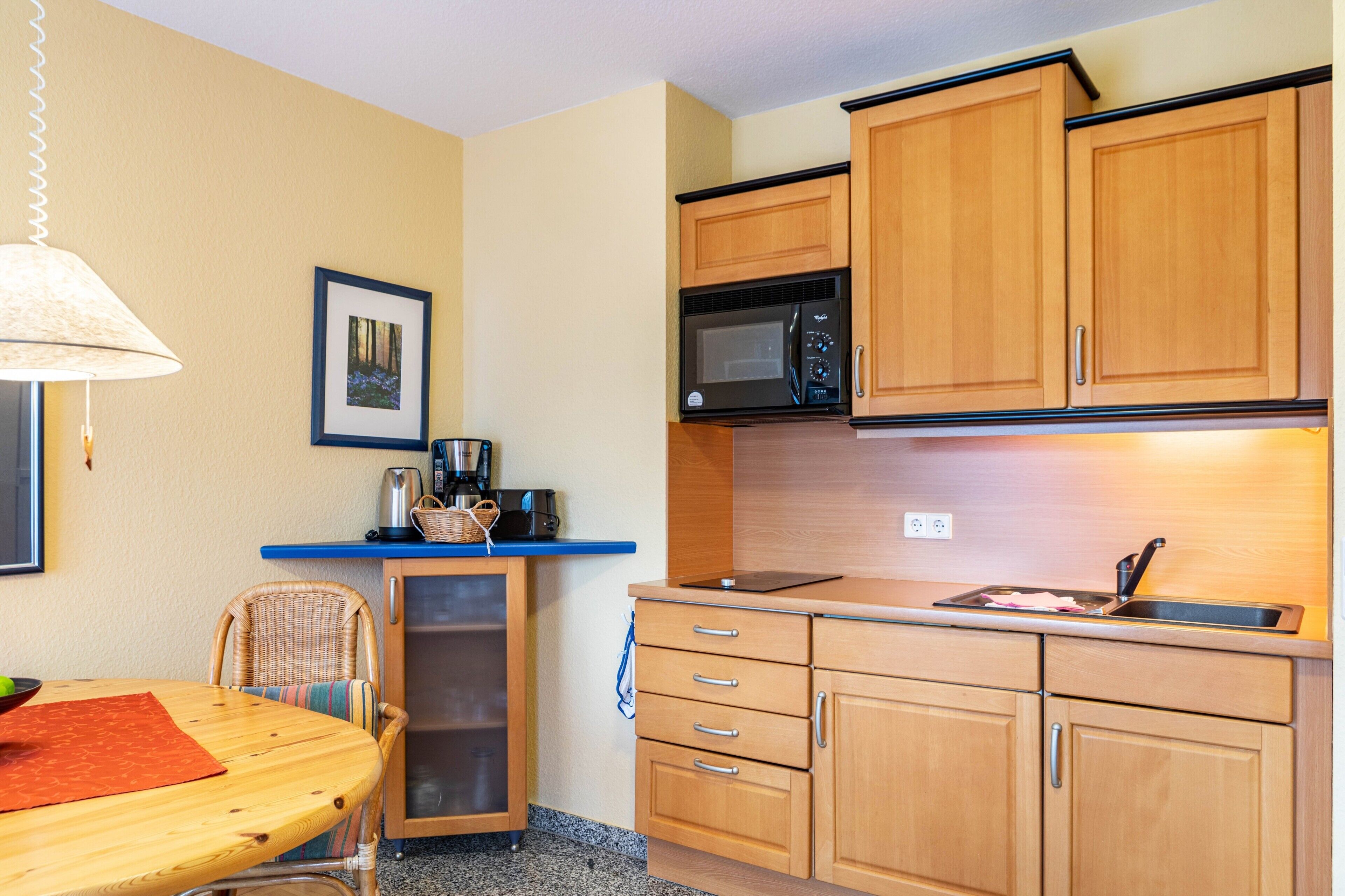 Cocina compacta con armarios de madera, microondas, lavamanos y una zona de comedor con mesa y silla.