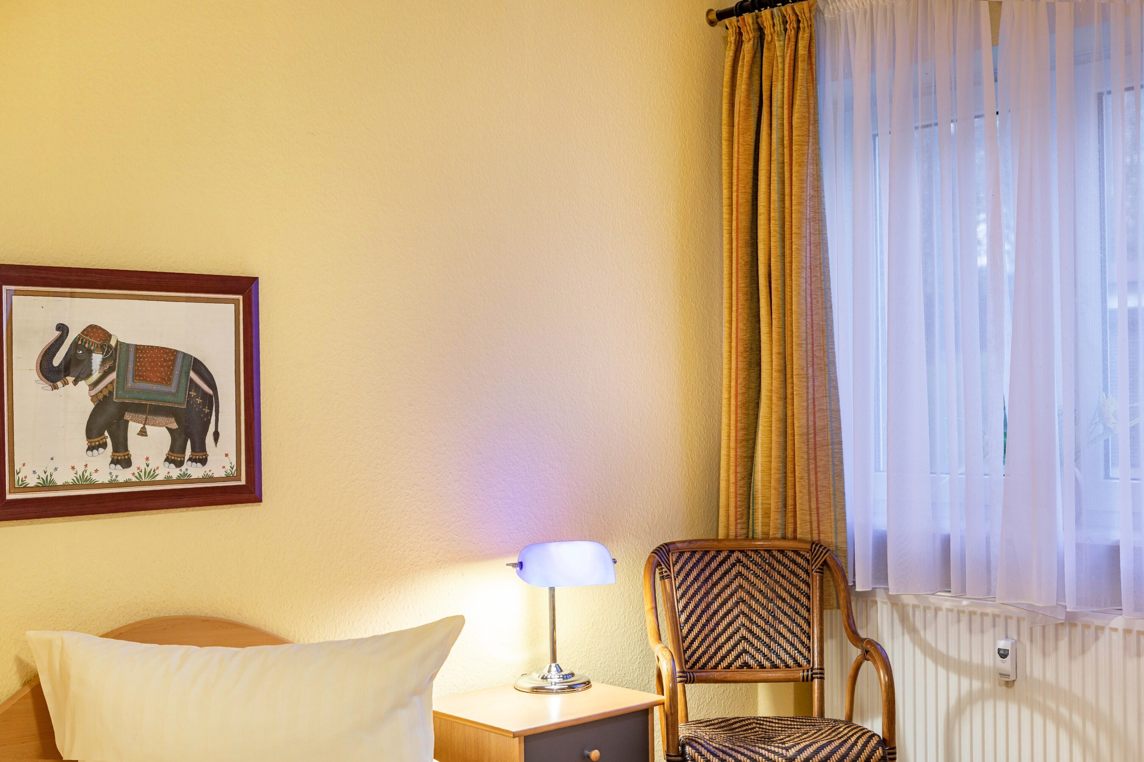 Una habitación de hotel con una cama, una silla, una mesita de noche con lámpara, un cuadro enmarcado de un elefante y una ventana con cortinas.