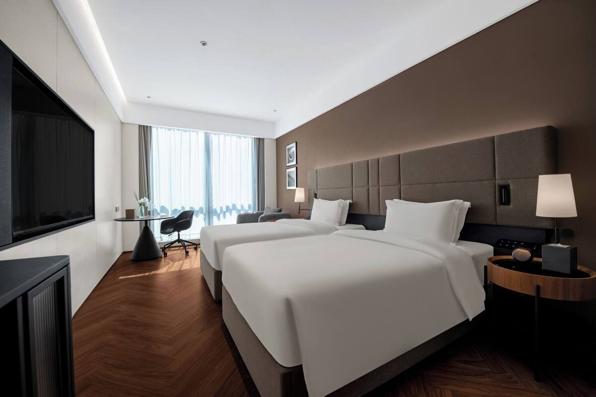 A hotel room with two beds, a desk, a chair, a TV, and a window with curtains.