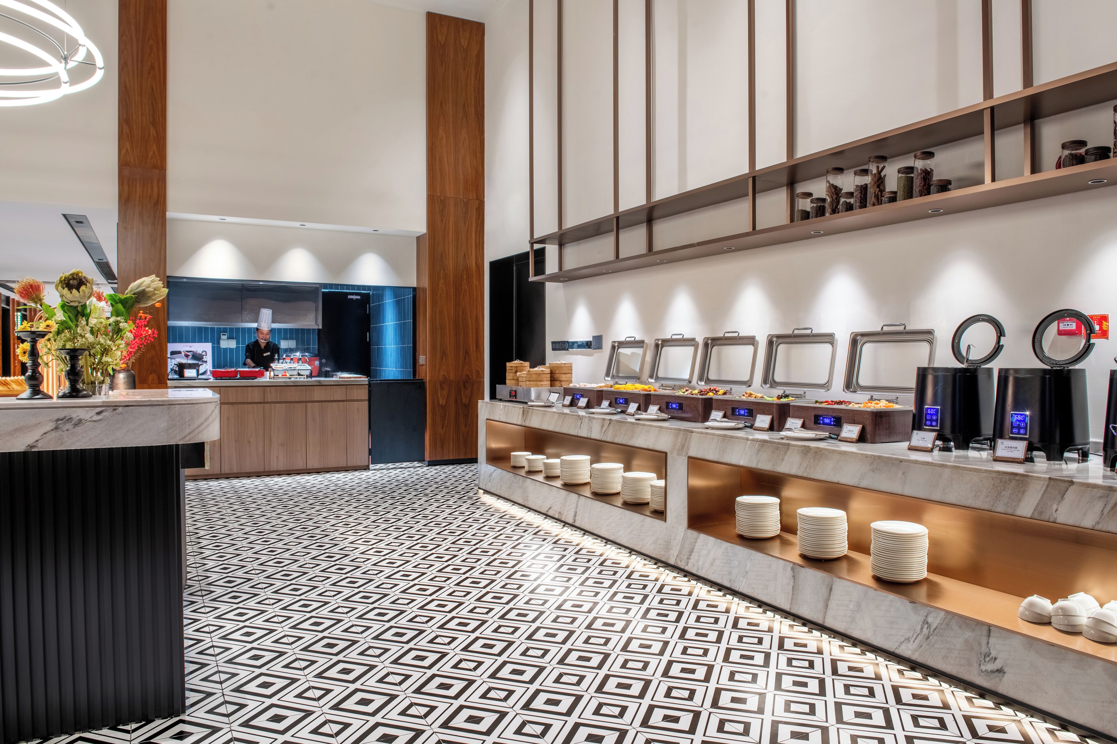 Um hall de hotel moderno com uma recepção de mármore, piso estampado e uma área de buffet com diversos alimentos e acompanhamentos.