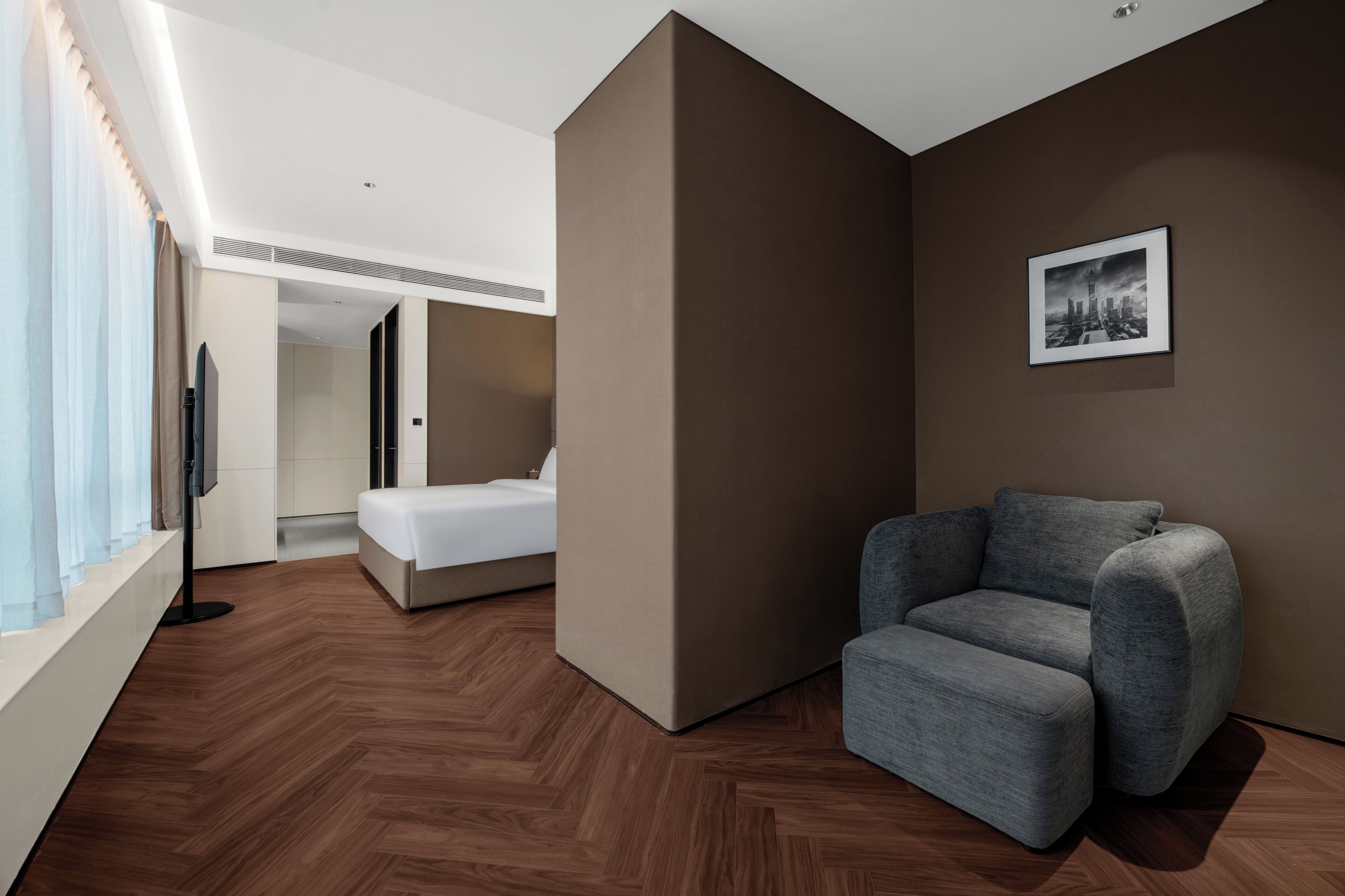 Une chambre d’hôtel moderne avec un lit, un canapé gris, un téléviseur à écran plat et un tableau représentant un paysage urbain accroché au mur.