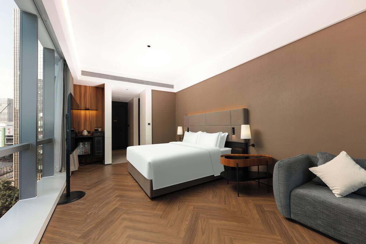A modern hotel room with a large bed, a sofa, a nightstand, and a view of the city.