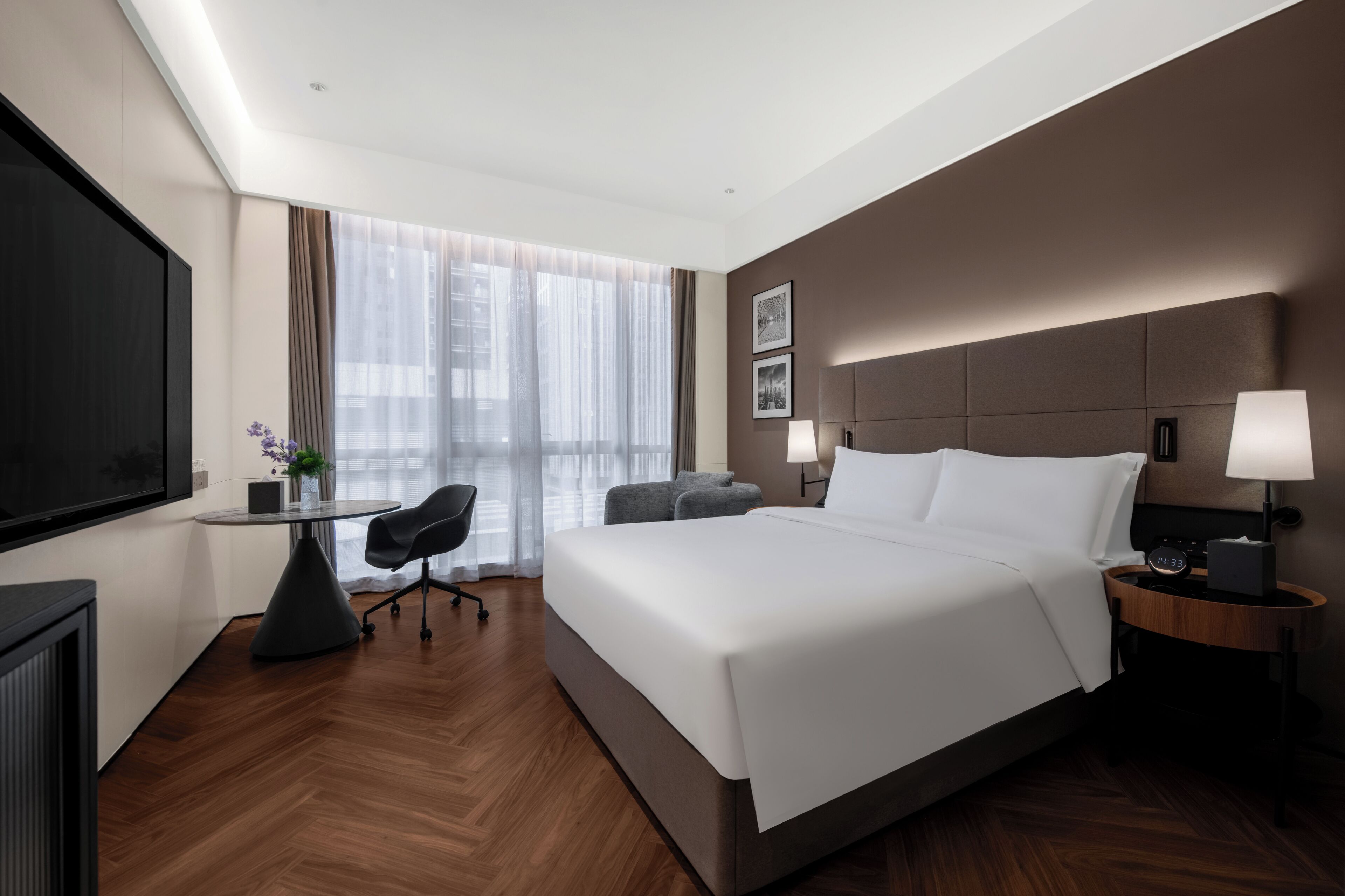 A modern hotel room with a large bed, a desk, a chair, a TV, and a window with sheer curtains.