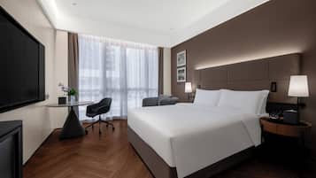 A modern hotel room with a large bed, a desk, a chair, a TV, and a window with sheer curtains.