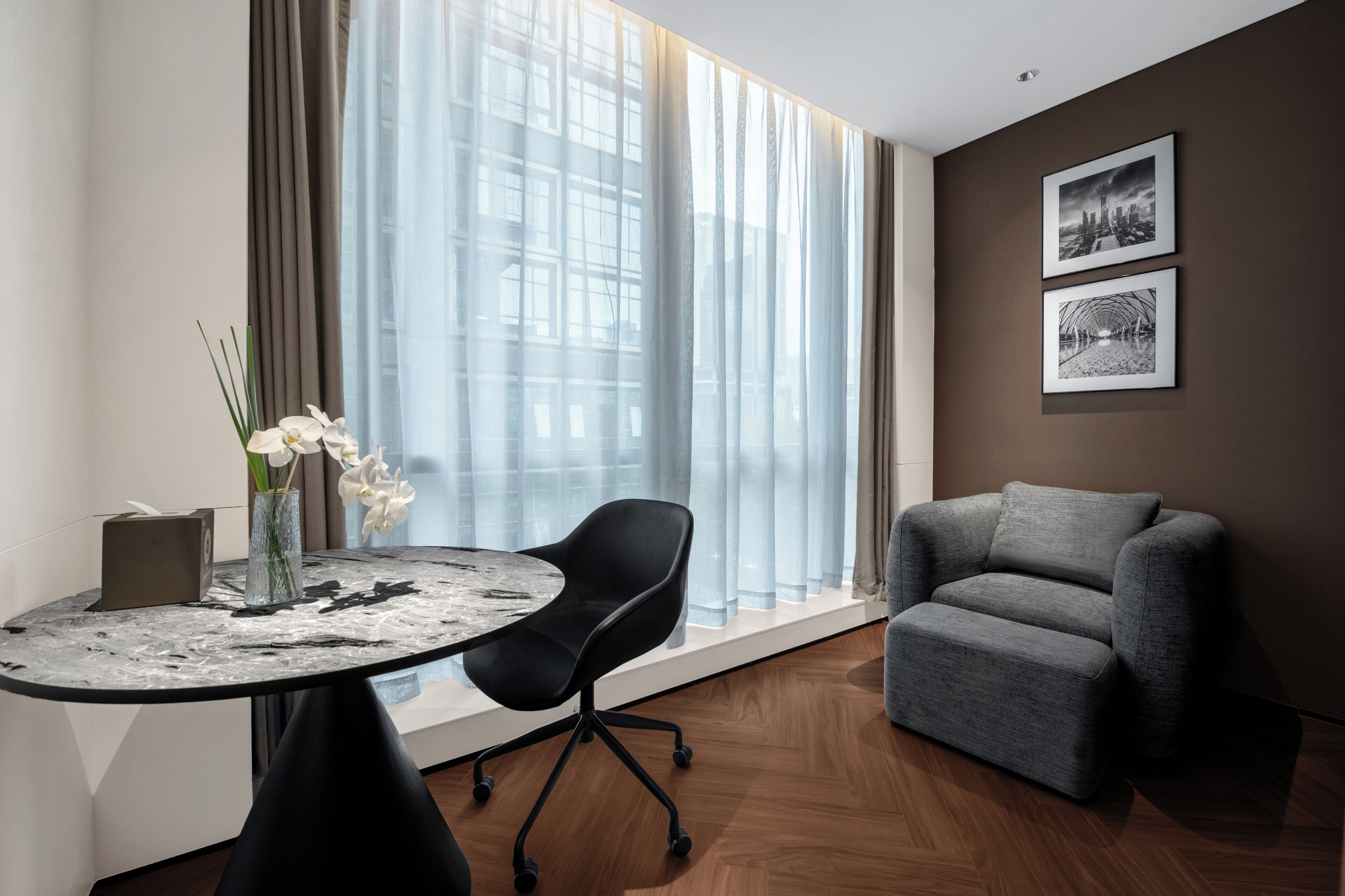 Ein modernes Zimmer mit rundem Tisch, schwarzem Bürostuhl, grauem Sofa, einer Vase mit weißen Blumen und zwei gerahmten Bildern an der Wand.