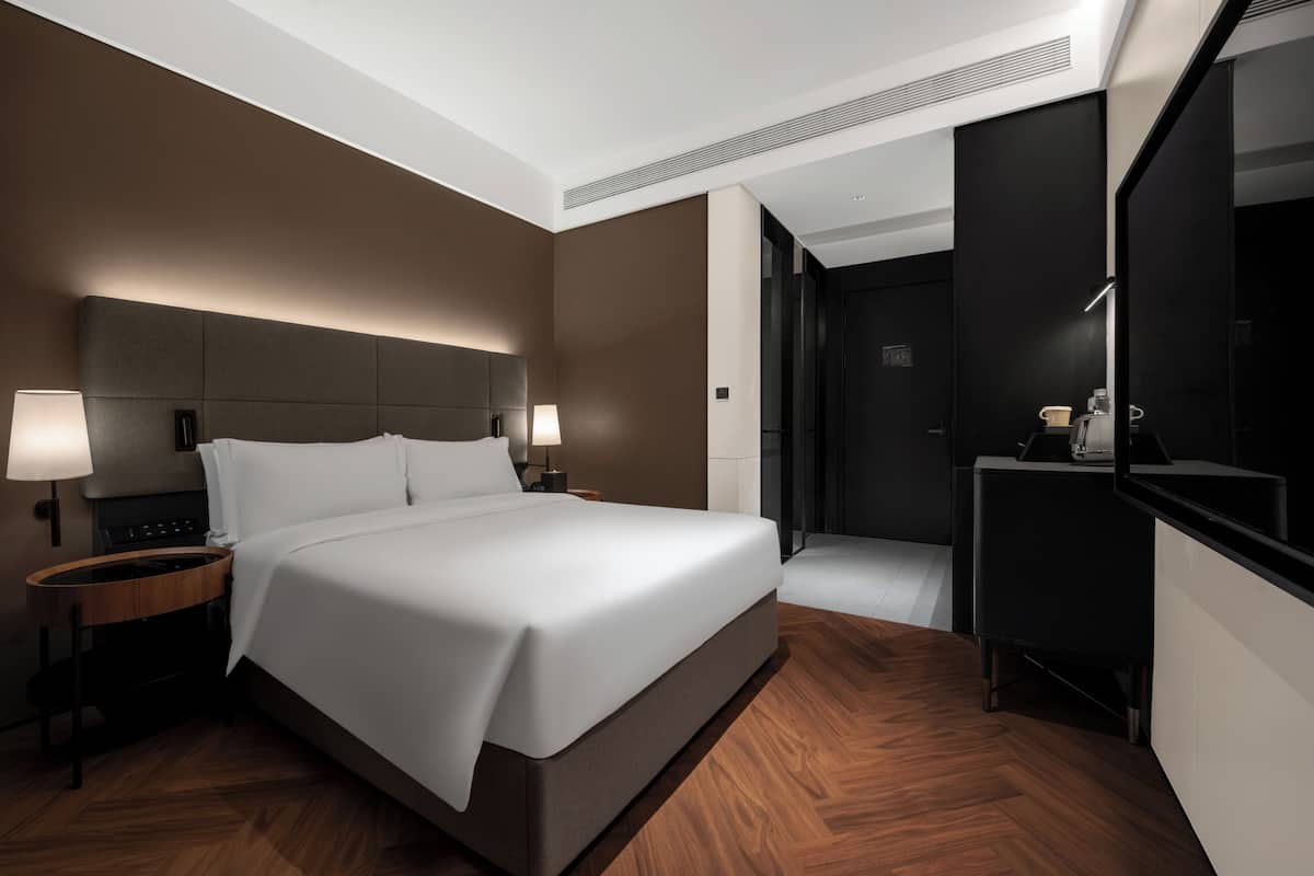A modern hotel room with a large bed, bedside lamps, a small table, a mirror, and a door leading to a bathroom.