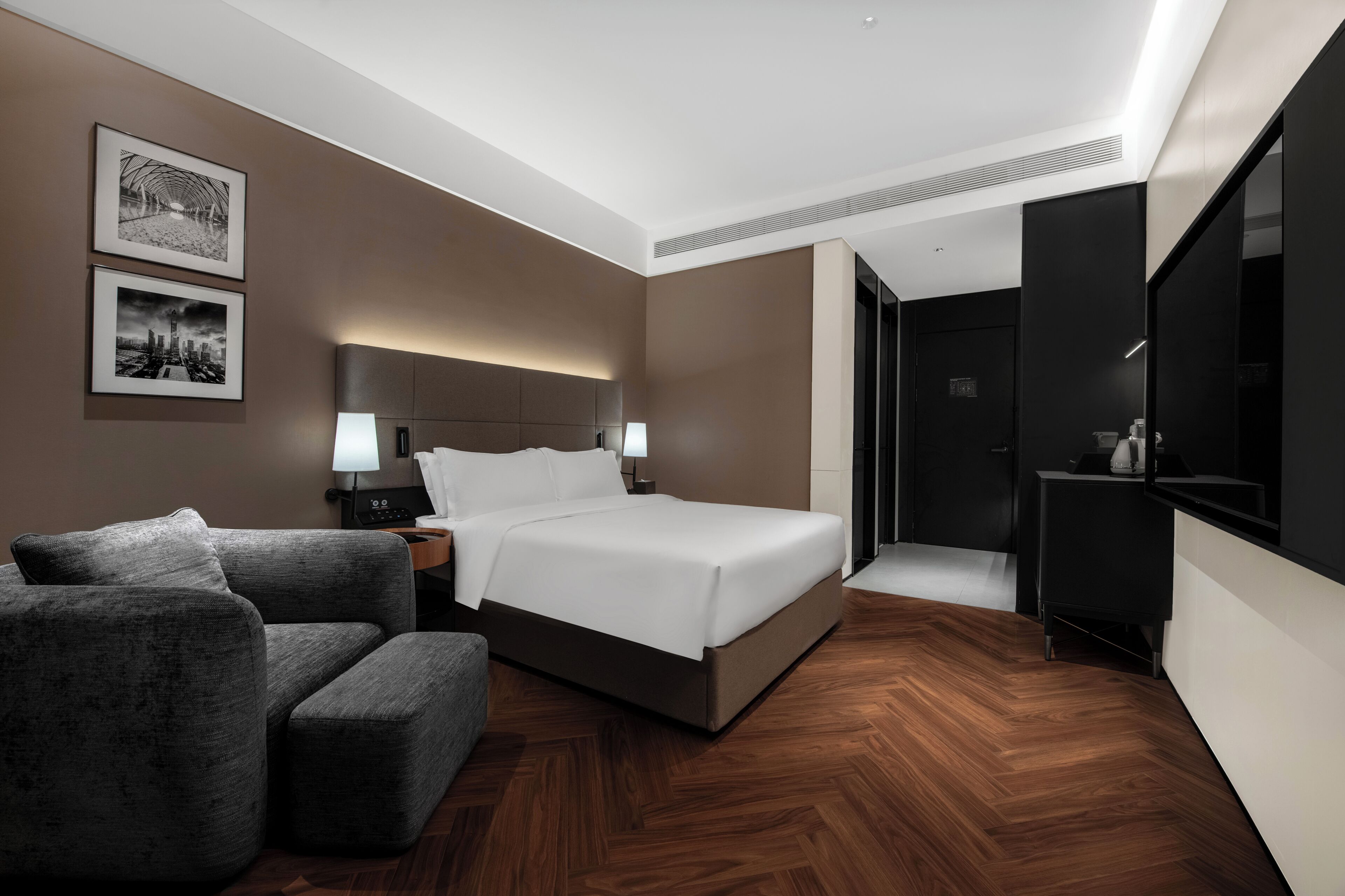 Ein modernes Hotelzimmer mit einem großen Bett, einem grauen Sofa, einem Schreibtisch mit Lampe und zwei gerahmten Bildern an der Wand.