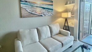 Ein Wohnzimmer mit einem beige farbenen Sofa, einem Glastisch, einem Gemälde mit Strandmotiv, einer Lampe und einer Tür mit strukturierter Wand.