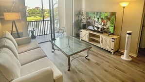 Ein Wohnzimmer mit Glastisch, weißem Sofa, Deckenventilator, Fernseher und Blick auf Pool und Grünfläche draußen.