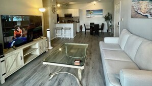 Ein Wohnzimmer mit Fernseher, einem Glastisch, einem weißen Sofa, einem Deckenventilator und einer Küche im Hintergrund.