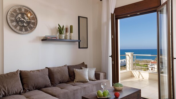Un salon avec un canapé, une table basse sur laquelle on trouve des fruits et un verre, une horloge, et une vue sur la mer par une porte vitrée.