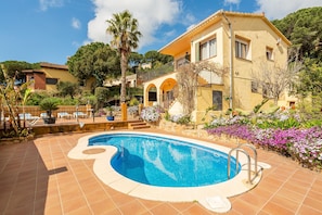 Una casa de dos pisos con piscina, rodeada de vegetación y flores.