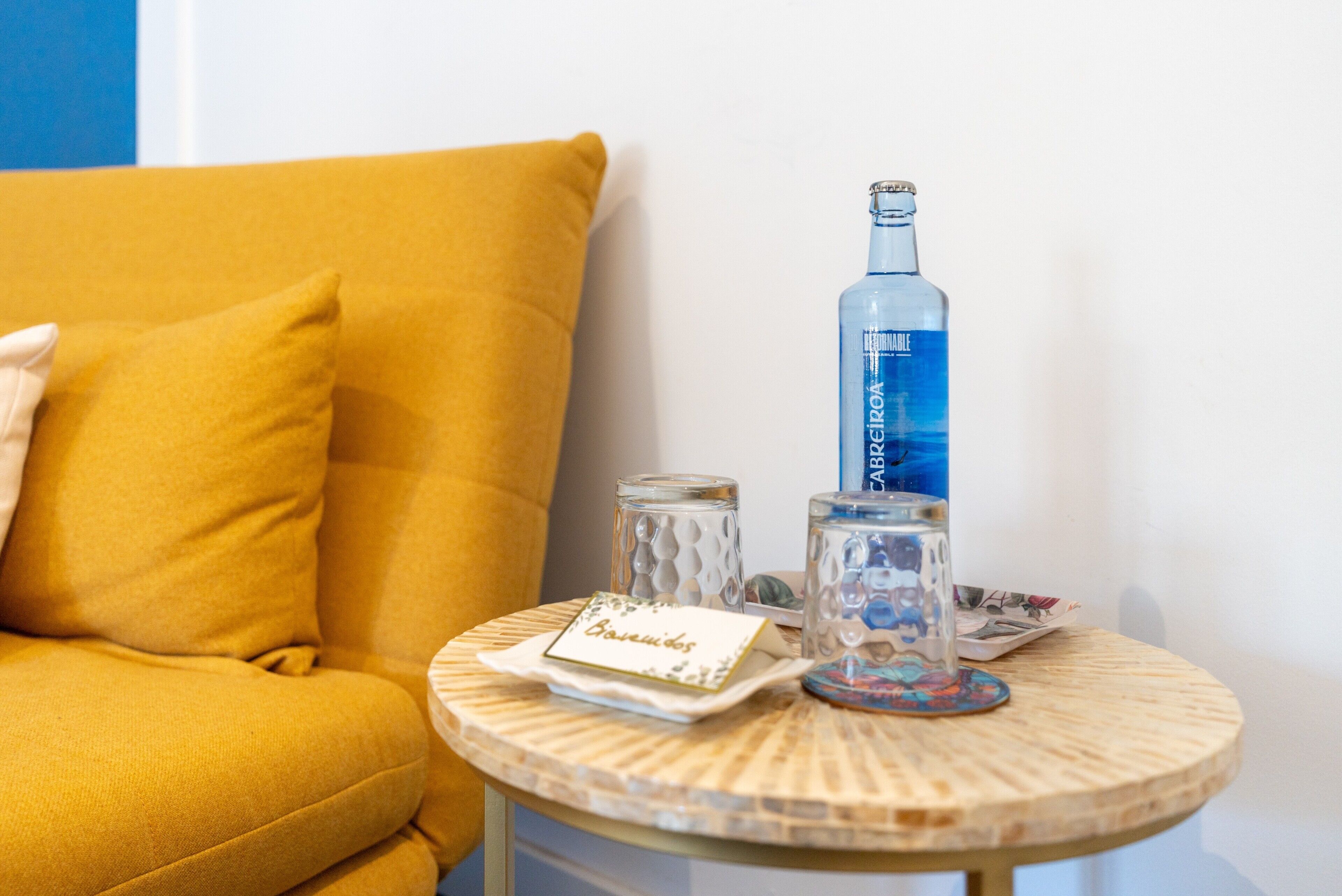 En gul sofa, et lille rundt bord med en flaske, to glas og et kort.