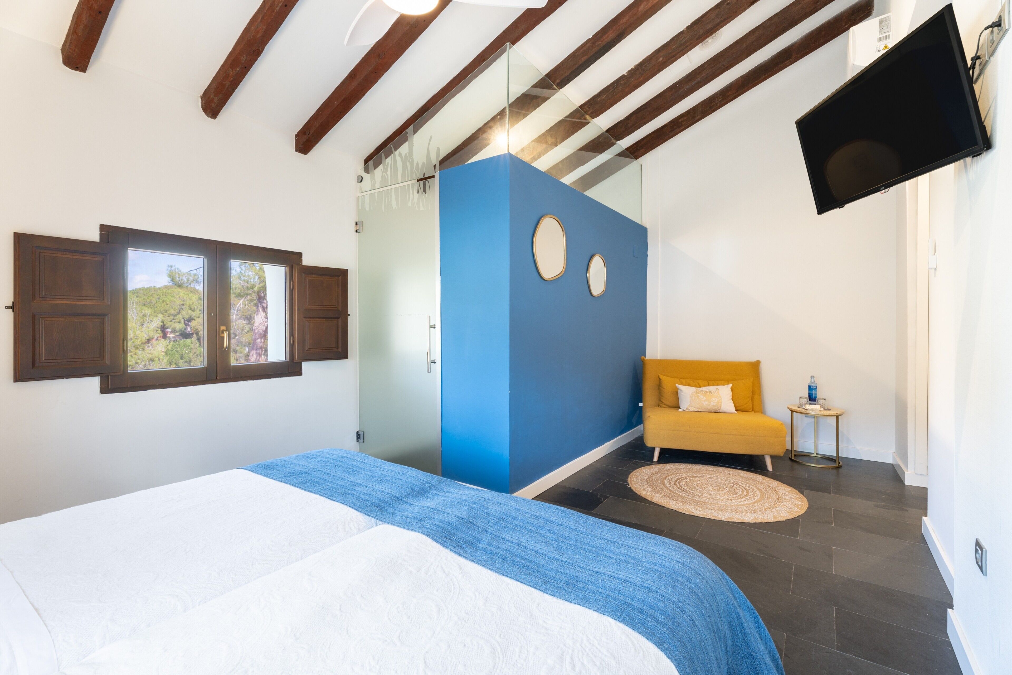 A bedroom with a bed, a window, a blue accent wall, a yellow armchair, and a flat-screen TV.