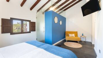 A bedroom with a bed, a window, a blue accent wall, a yellow armchair, and a flat-screen TV.