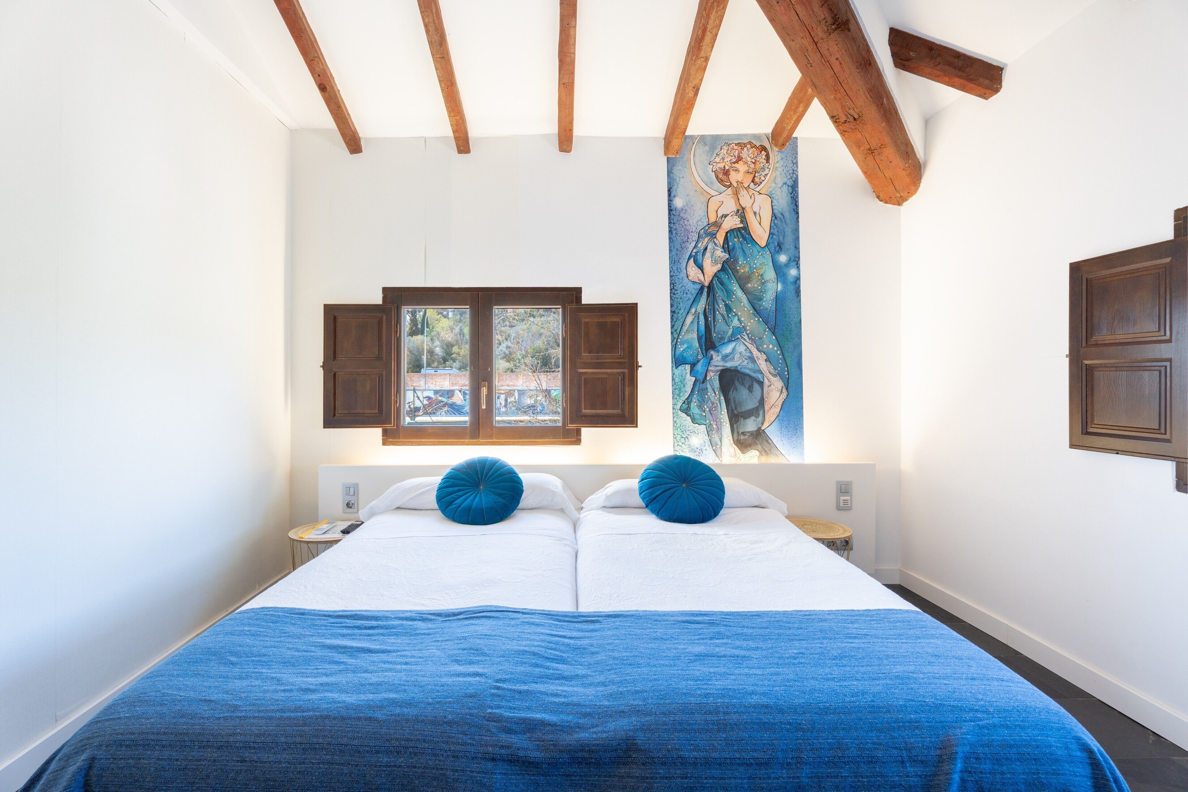 Een tweepersoonsbed met een blauwe deken, houten balken, een raam met gordijnen en een aan de muur hangend schilderij.