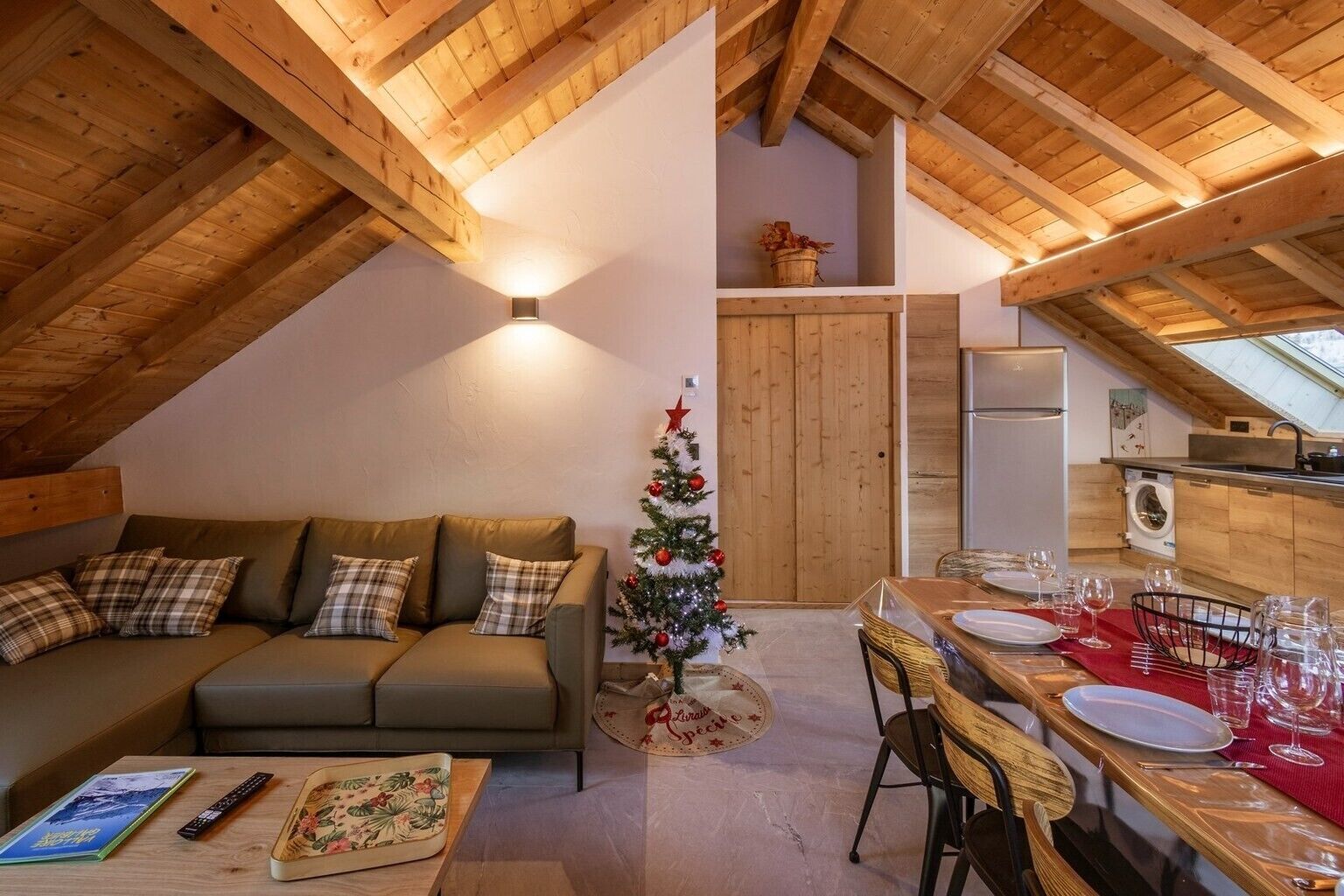 Ein gemütliches Loft mit Holzbalkendecke, einer Couch mit Karokissen, einem kleinen Weihnachtsbaum, einem Esstisch für vier Personen und einer Waschmaschine im Hintergrund.