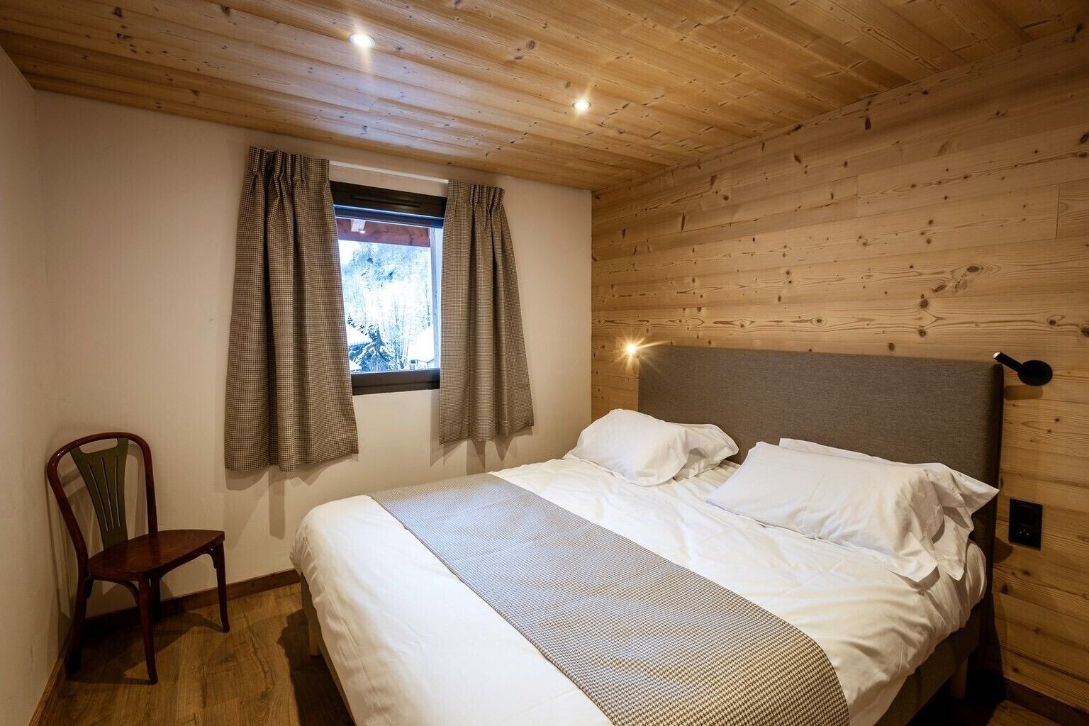 Une chambre avec un plafond en bois, un lit recouvert de draps blancs, une fenêtre avec des rideaux et une chaise.