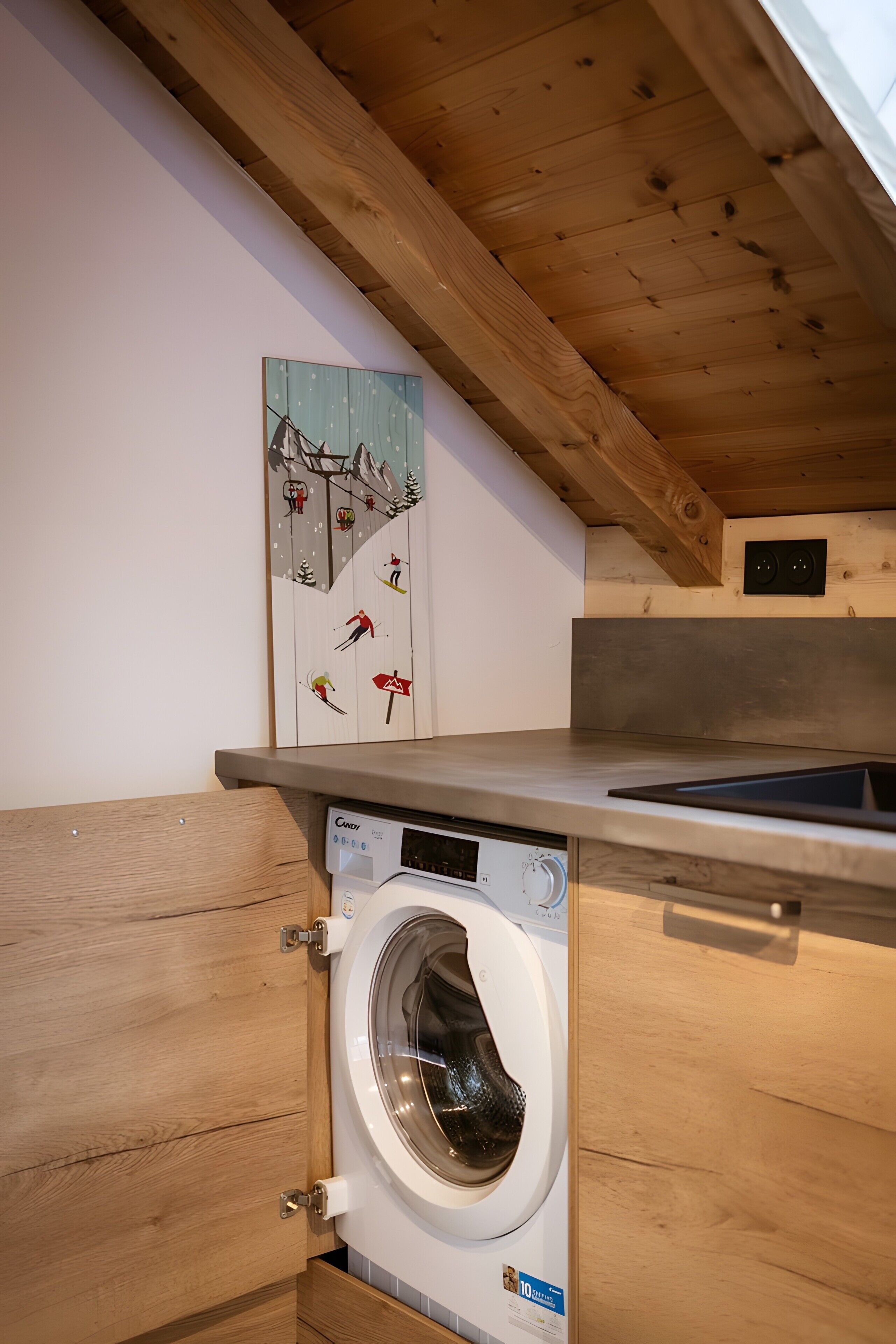 A compact kitchen with a built-in washing machine, wooden ceiling beams, and a wall-mounted artwork depicting skiers.