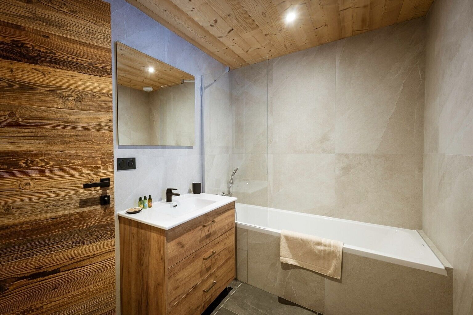 A modern bathroom with a wooden vanity, a large mirror, a bathtub, and tiled walls.