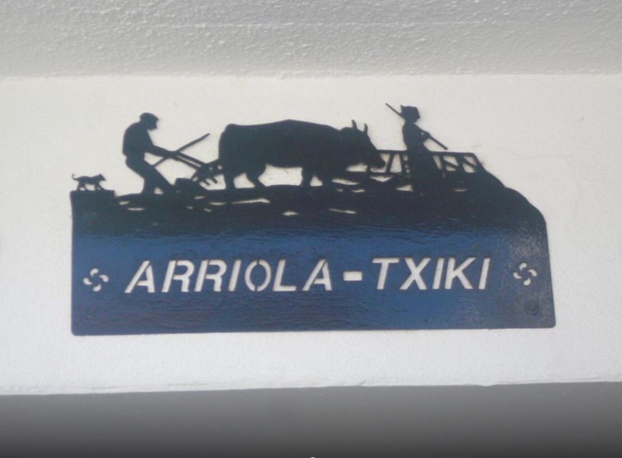 Silueta de una persona con un perro y una vaca en un vehículo, y el texto 'ARRIOLA-TXIKI'.